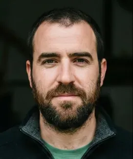 Hombre con barba y cabello corto, mirando a cámara con expresión seria, fondo desenfocado.